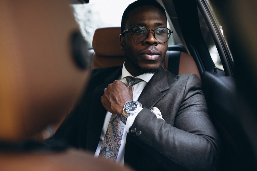 Man adjusts tie while sitting in a car, wearing a dark suit and glasses, with a watch visible on his wrist.
