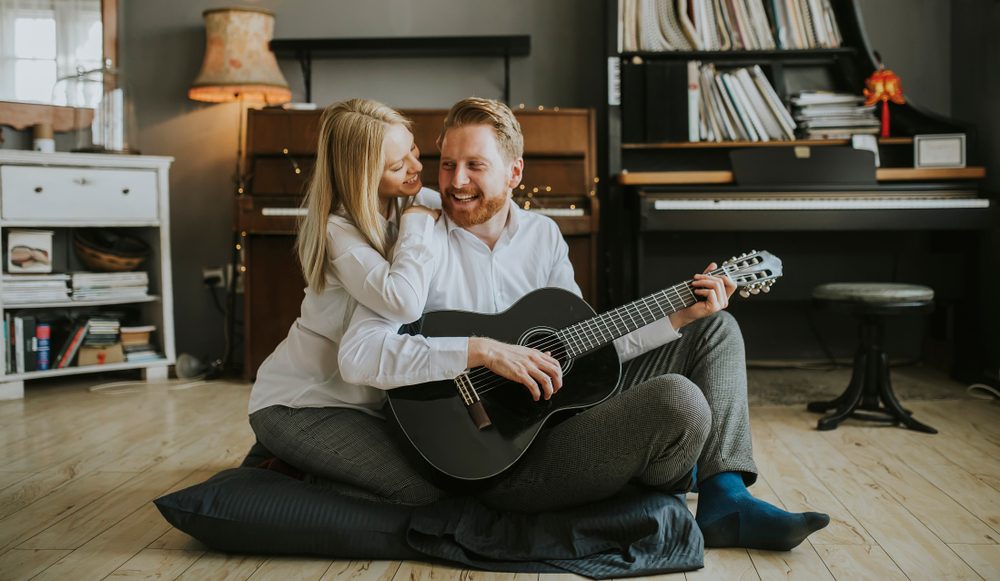Man plays guitar sitting on cushions, woman leans affectionately on his shoulder; surrounded by a cozy room with a piano and bookshelves.