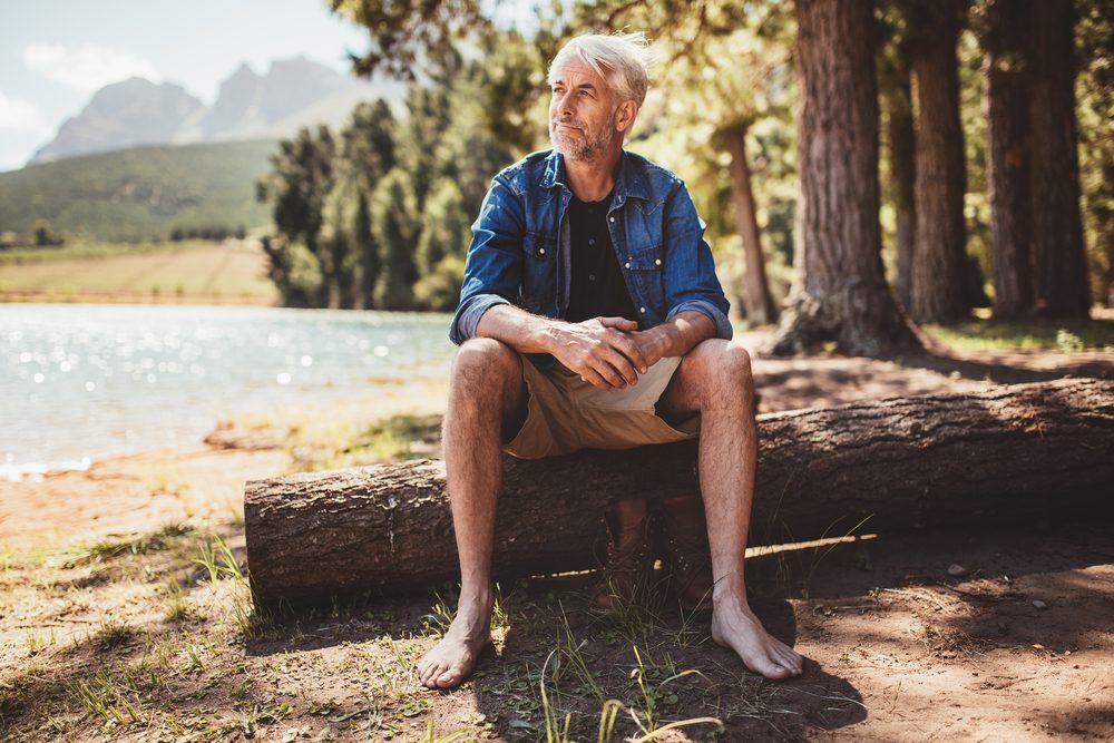 Man sits on a fallen log, gazing into the distance by a lakeside surrounded by trees under clear skies.