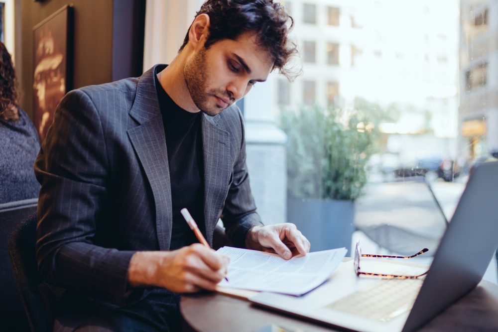 Man writing notes in a notebook at a cafe table with a laptop, large window, and city street view in the background.