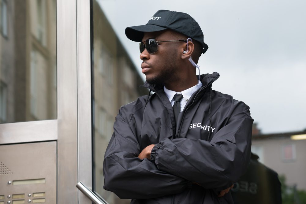 Security guard stands with arms crossed, wearing sunglasses and uniform, outside a building with glass doors.