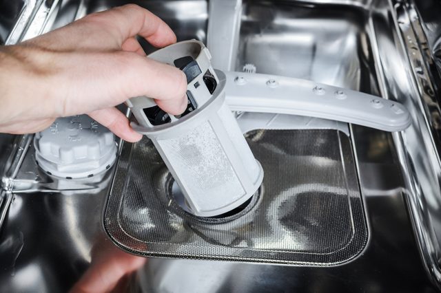 Hand removes the filter from a dishwasher's metal interior, surrounded by spray arms and a clean, stainless environment.