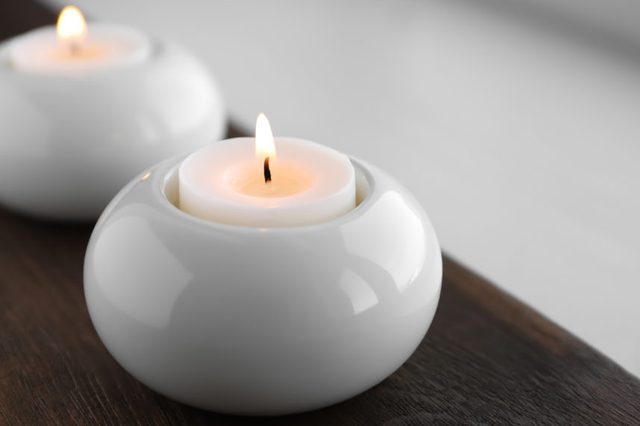 A white candle holder supports a lit candle, placed on a dark wooden surface, with another blurred candle holder in the background.