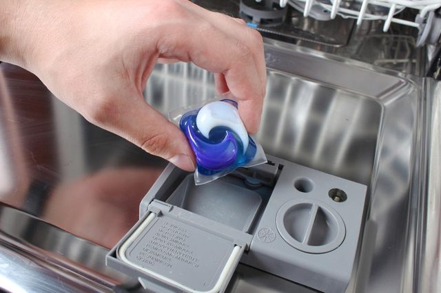 A hand places a detergent pod into an open dishwasher detergent compartment.