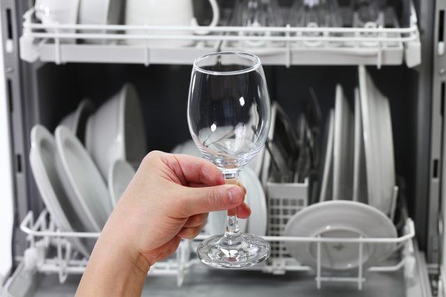 Hand holds wine glass, removes it from dishwasher filled with clean, organized dishes and utensils.