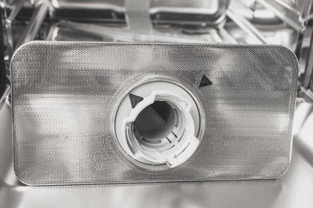 A silver mesh dishwasher filter rests inside a metallic dishwasher, surrounded by reflective surfaces.