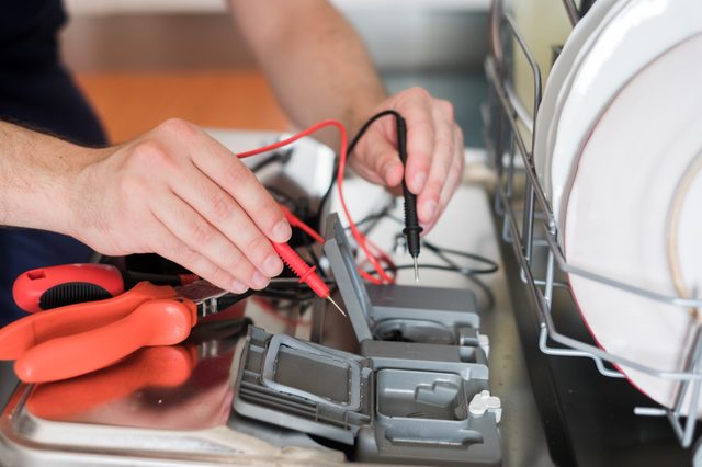 Hands test dishwasher components with multimeter probes beside open detergent compartment in kitchen setting.