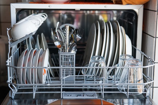Dishwasher holding clean plates, glasses, and silverware, is open in a tiled kitchen space.