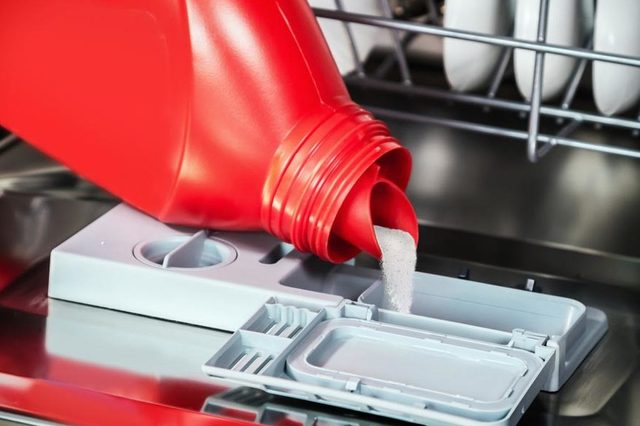 Red bottle pouring powder into dishwasher detergent compartment, surrounded by open dishwasher door and dish rack with plates.
