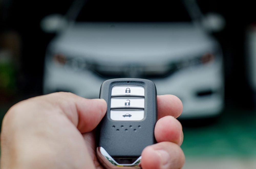 Hand holds a car key fob, poised to unlock, with a blurred white car in the background.