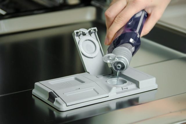 Hand pours liquid detergent into dishwasher dispenser, located on a stainless steel surface.