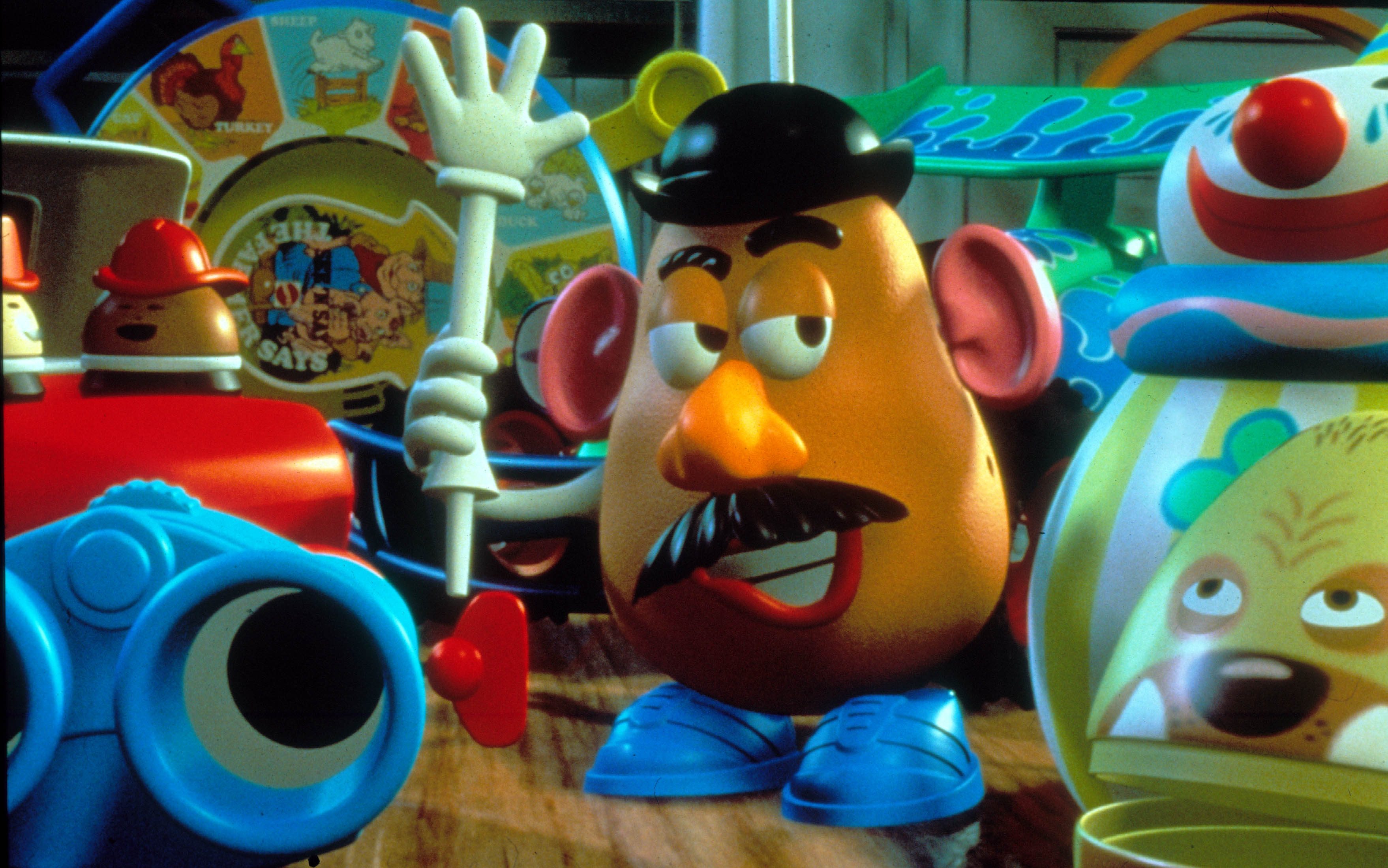 Toy potato character holds a white arm amidst colorful, playful objects in a toy room setting.