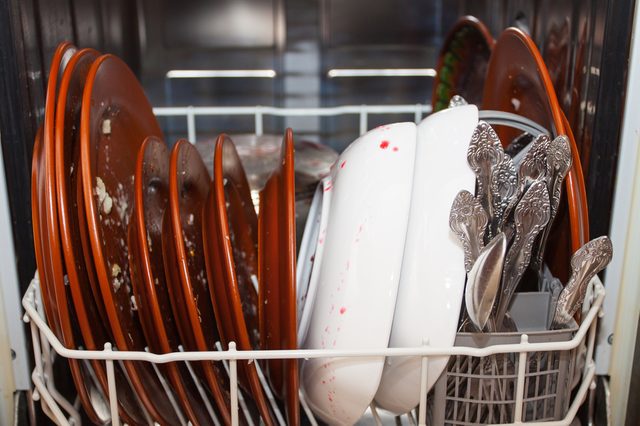 Plates and utensils rest in a dishwasher rack, covered with food residue, awaiting cleaning.