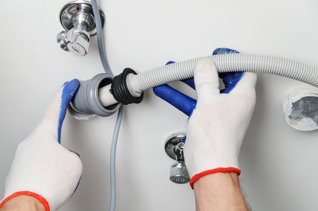 Hands connect flexible gray hose to water outlet; wearing protective gloves; white wall background.