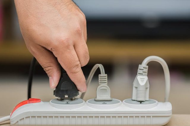 Hand inserts black plug into power strip with multiple connected cables, placed on a table in an indoor setting.