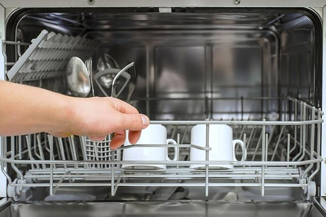 A hand closes a dishwasher rack containing two white mugs and assorted cutlery in a kitchen setting.