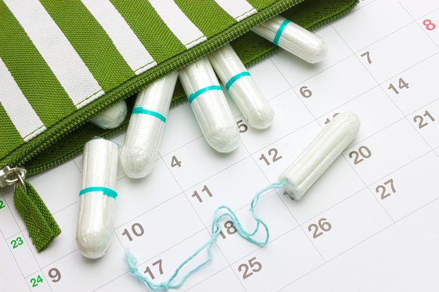 Tampons spilling from a zippered green striped pouch onto a calendar, dates visible, suggesting menstrual period tracking.