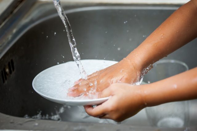 Hands wash a white plate under running water in a stainless steel sink.