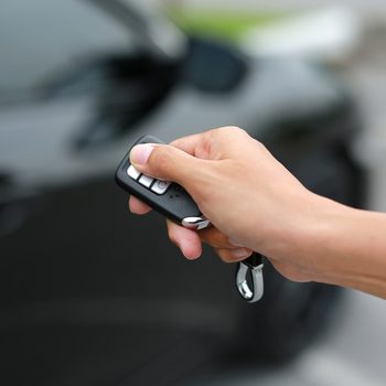 Hand holding a car remote, pressing a button, in front of a parked black car outdoors.