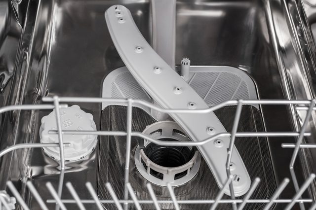 Dishwasher interior showing rotating arm and filter in a metallic environment, surrounded by wire rack compartments.