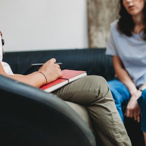 A person holds a pen over a red notebook, engaged in conversation on black chairs in a counseling setting.