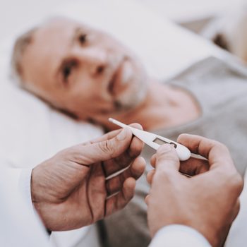 Hands holding a digital thermometer, displaying temperature, over a hospitalized man lying in bed with a concerned expression.