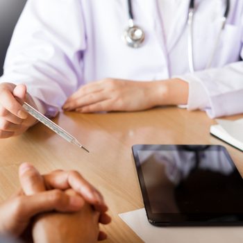 Doctor holds a thermometer, sitting at a desk with a patient, notepad, and tablet.
