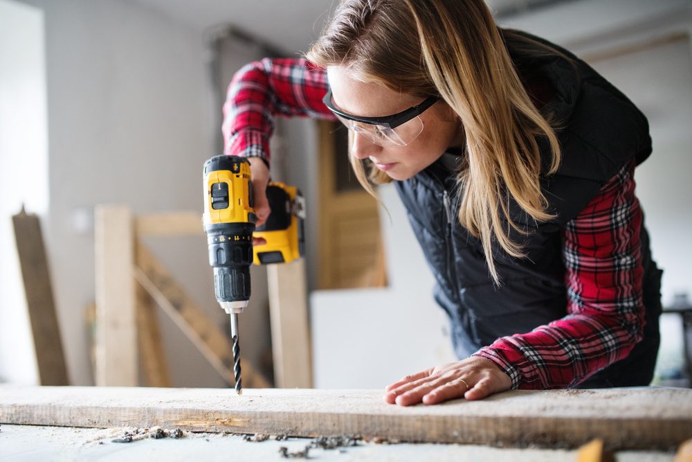 Person uses a power drill on a wooden plank, wearing protective goggles in a workshop setting.