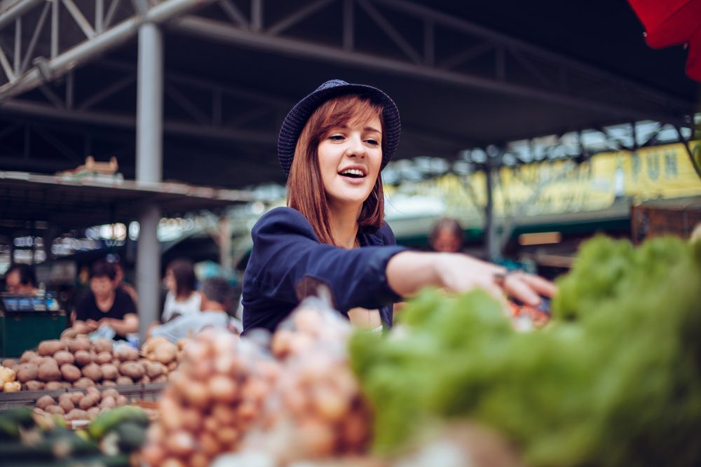 A woman selects produce at an outdoor market, surrounded by various vegetables and other shoppers under a metal canopy.