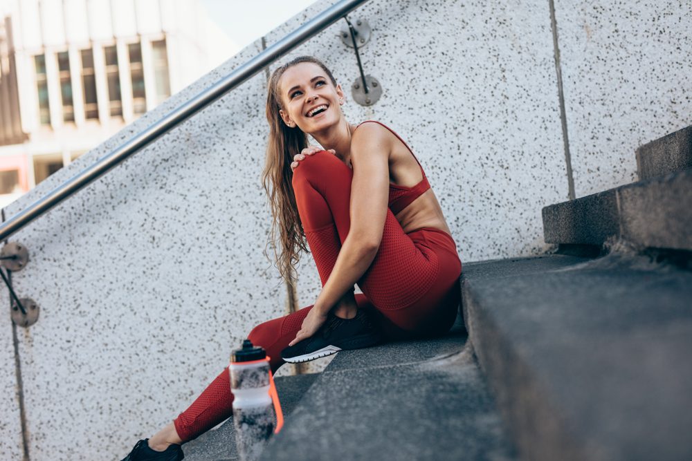 A person in red activewear smiles, sitting on concrete stairs outdoors, beside a water bottle.