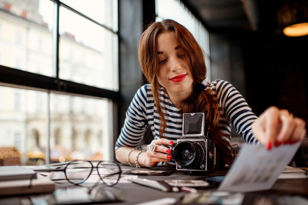 A woman examines photographs, holding a vintage camera, in a well-lit room with large windows and a cluttered desk.