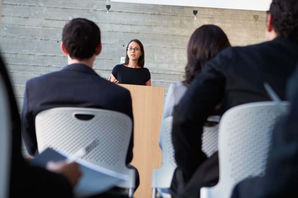 A person speaks at a wooden podium, addressing an audience in a modern conference room with a concrete wall background.