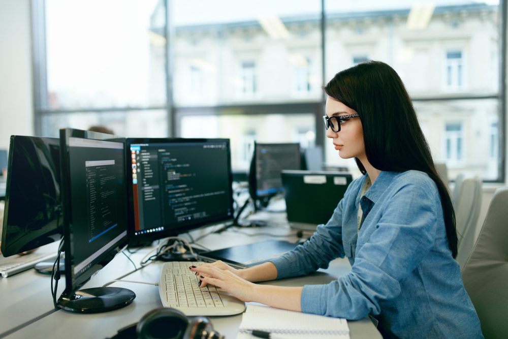 A person types on a keyboard, coding on dual monitors in an open-plan office with large windows.
