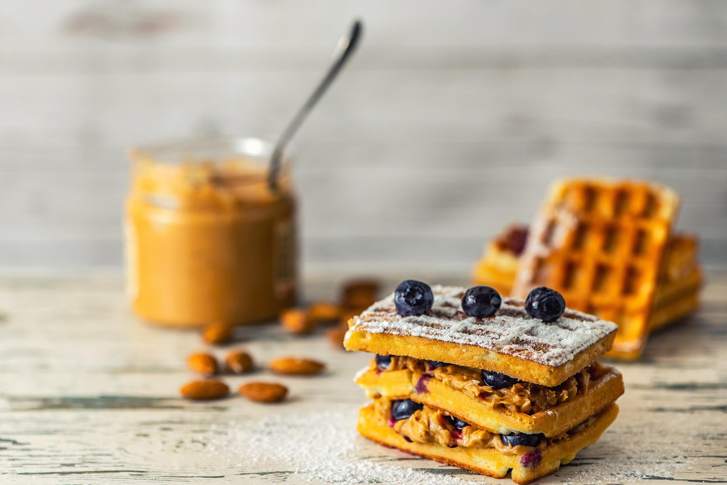 Waffles layered with peanut butter and blueberries, dusted with powdered sugar, rest on a wooden surface. Jar of peanut butter and almonds nearby.