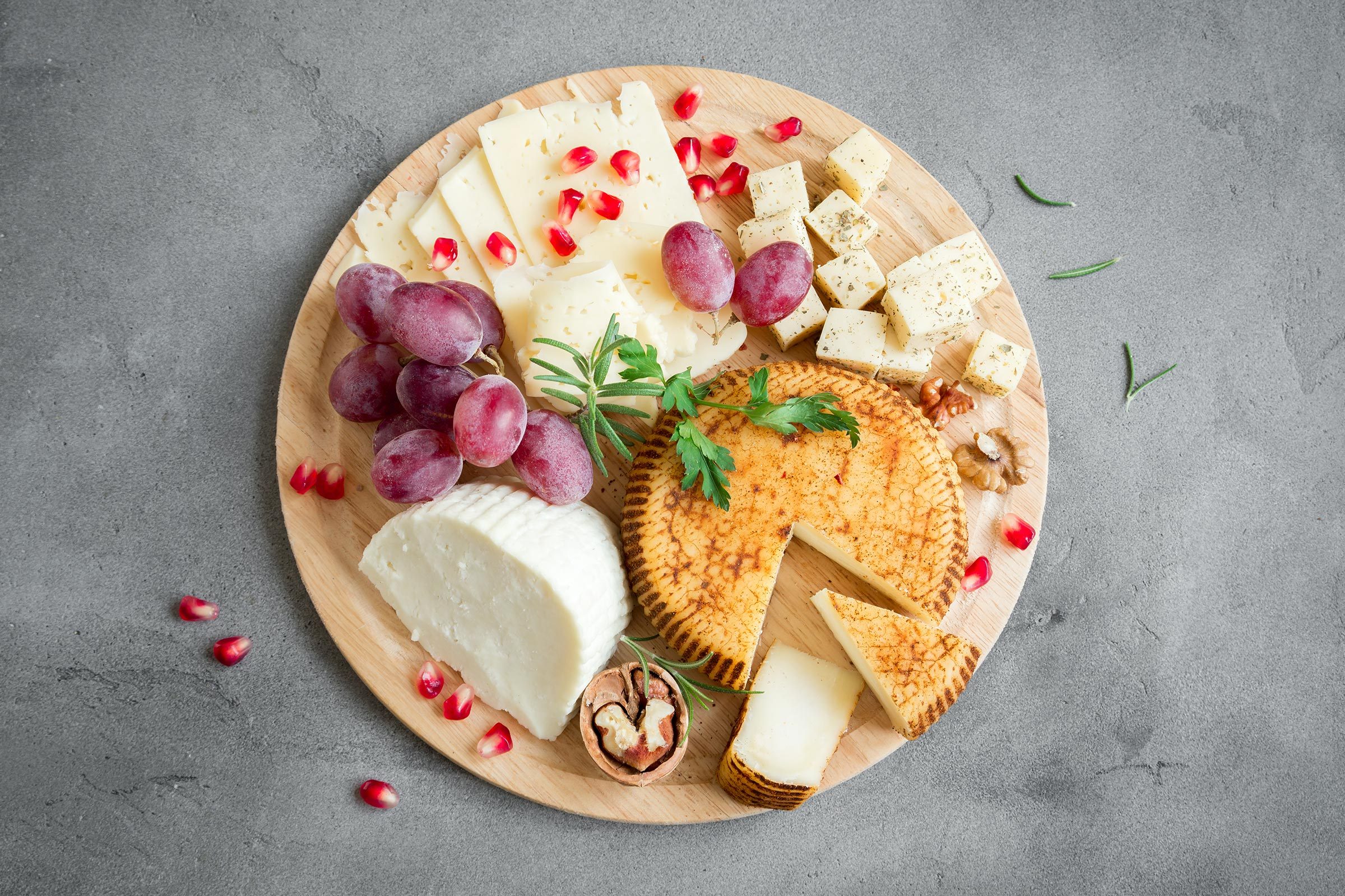 Cheese platter displays assorted cheeses, grapes, and nuts on a wooden board, garnished with pomegranate seeds and fresh herbs, against a gray surface backdrop.