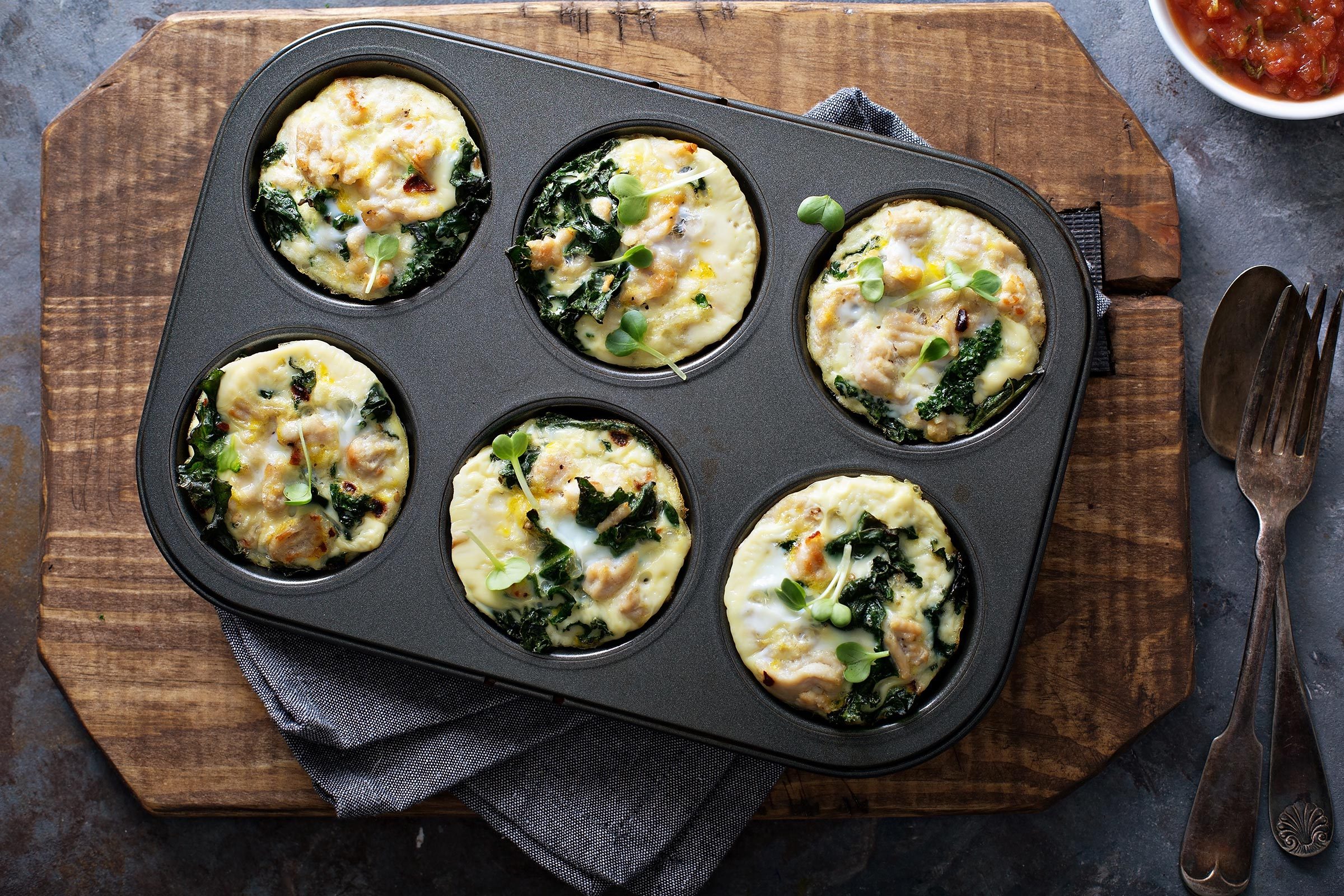 Muffin tin holds six baked egg cups with greens on a wooden board; cutlery and salsa dip nearby.