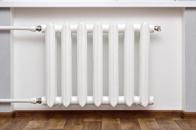 White radiator hanging on wall, situated above wooden floor, connected by pipes in a modern, minimalistic interior setting.