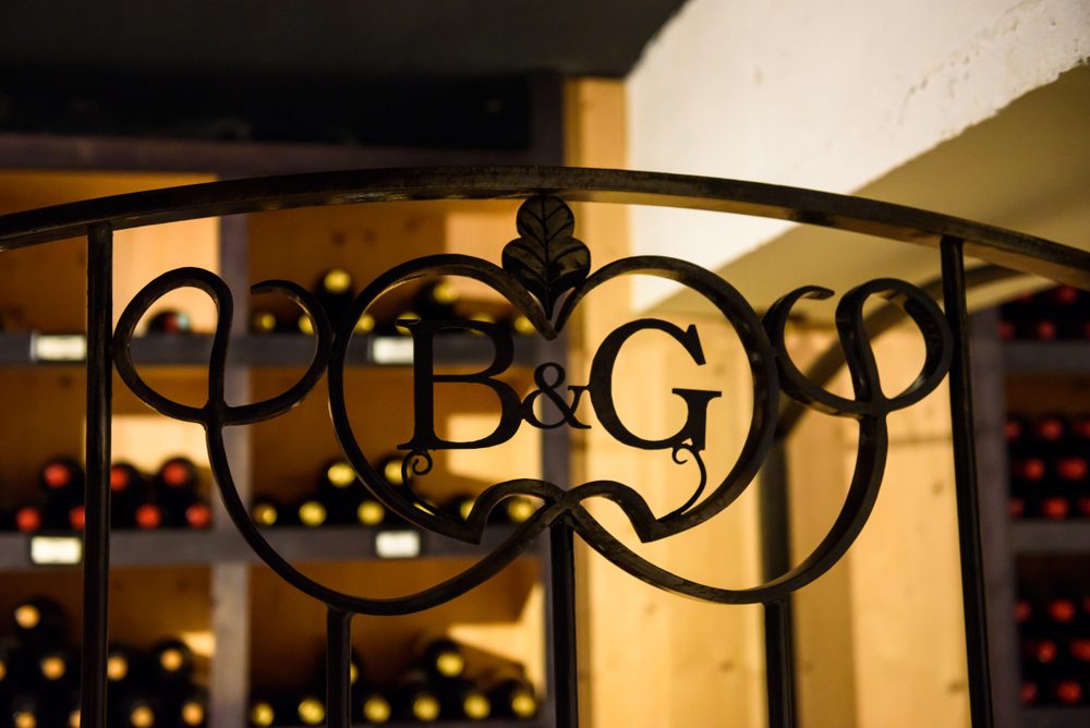 Ornate iron gate features "B&G" emblem, situated in a wine cellar with shelves of neatly arranged wine bottles as the background.