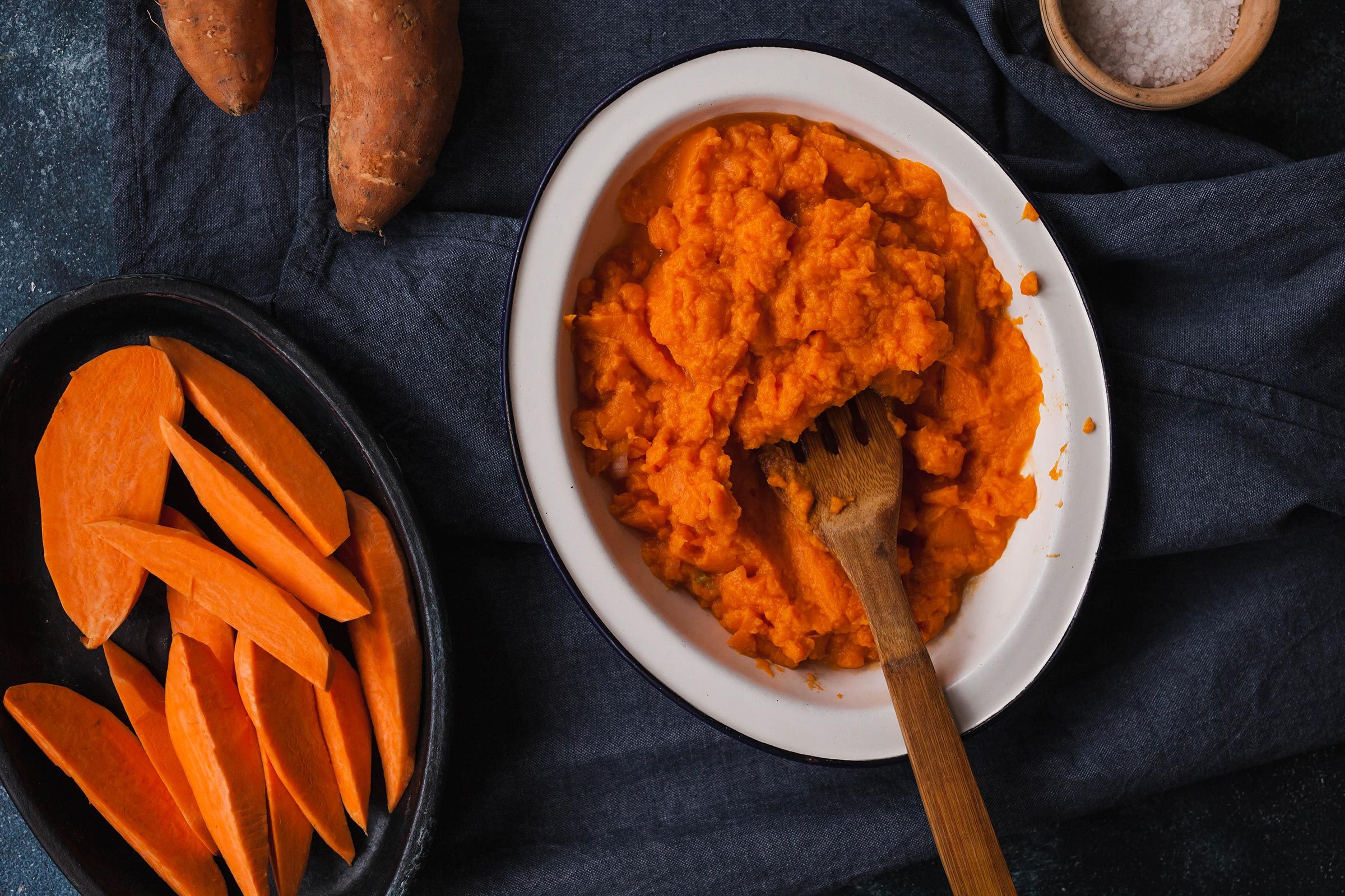 Mashed sweet potatoes sit in a white bowl with a wooden spoon; sliced pieces and whole sweet potatoes beside on a dark cloth.