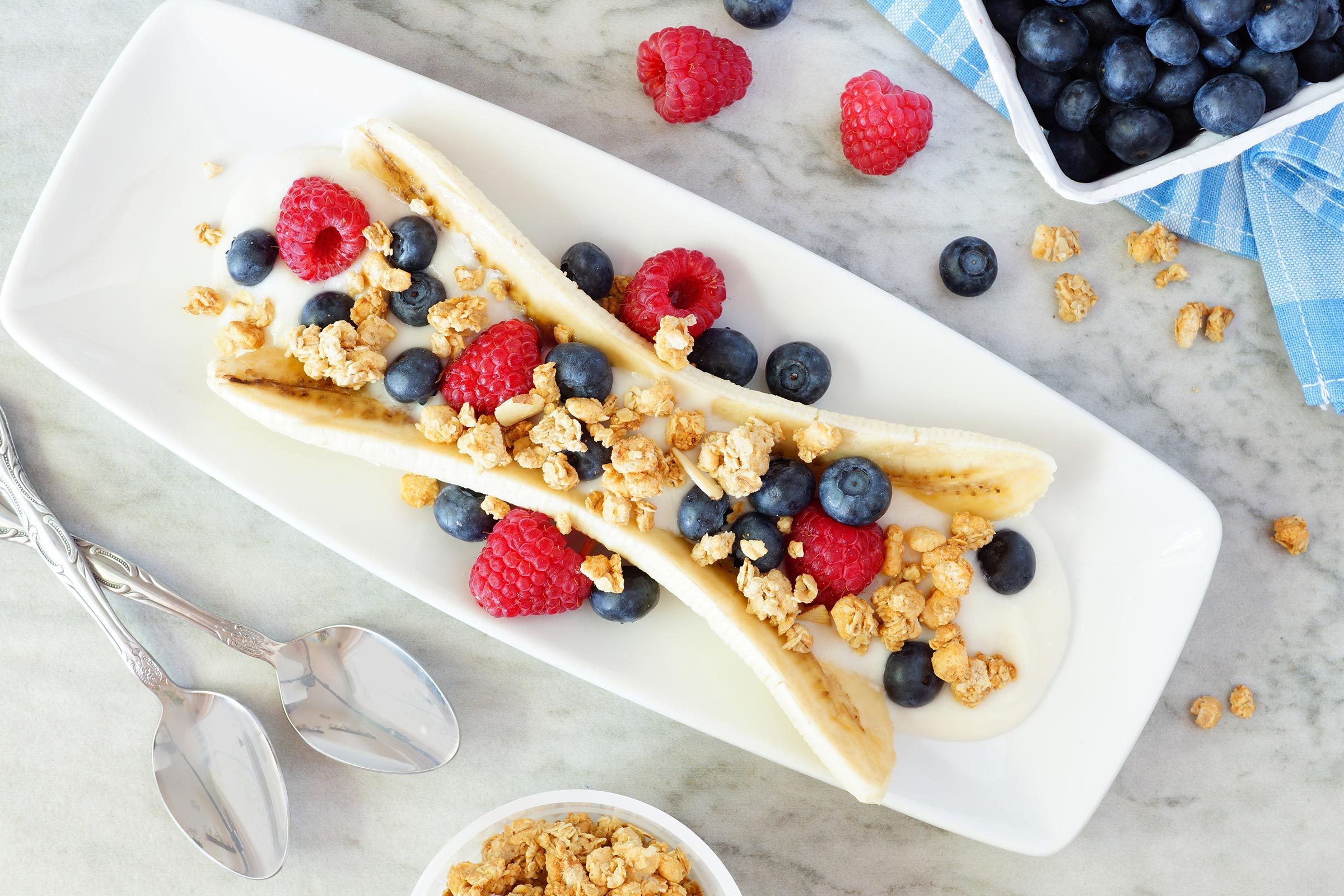 Banana split topped with raspberries, blueberries, and granola, placed on a white rectangular plate on a marble surface with spoons nearby.
