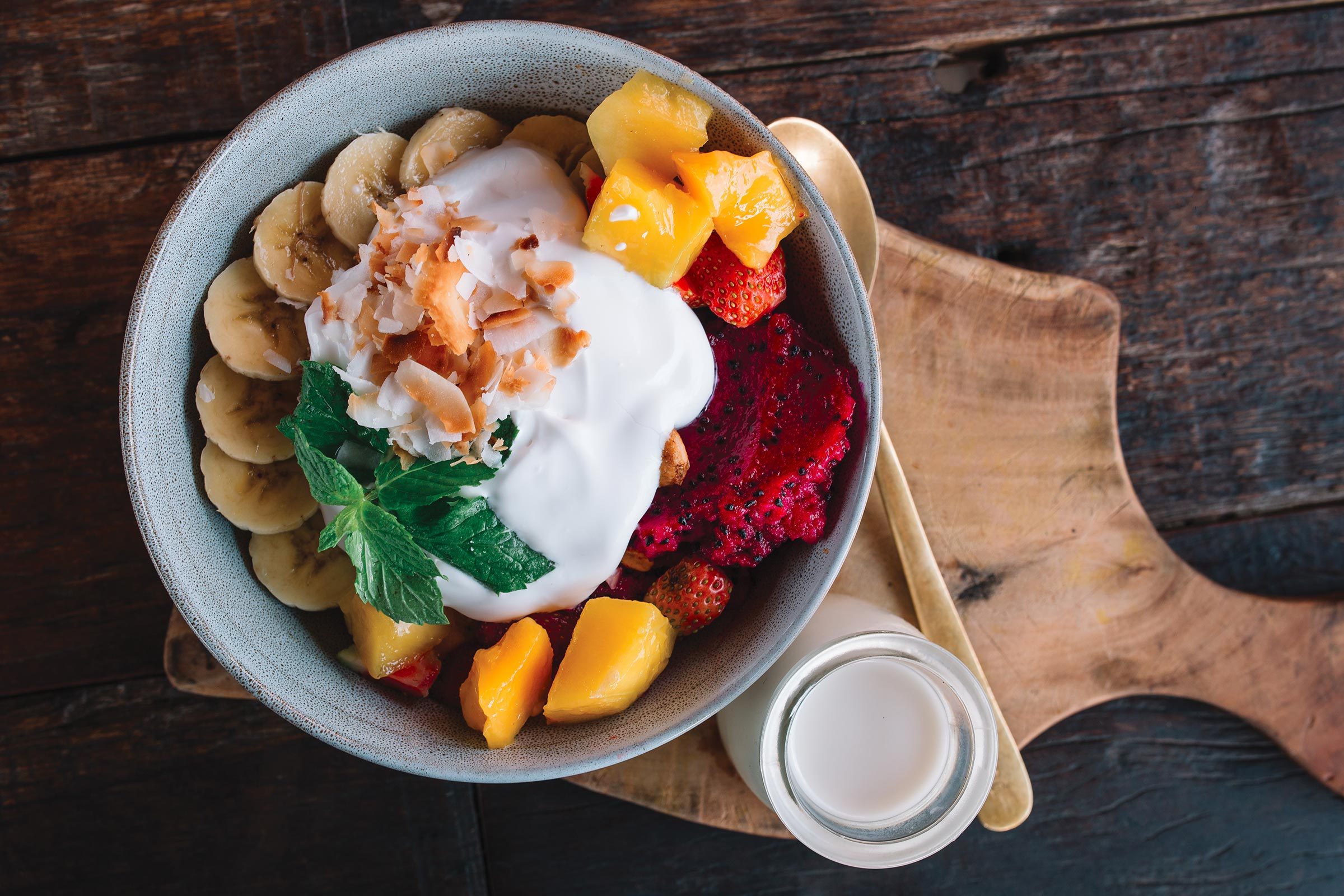 A bowl holds sliced bananas, yogurt, mango, and berries, garnished with mint on a wooden board beside a small bottle of milk.