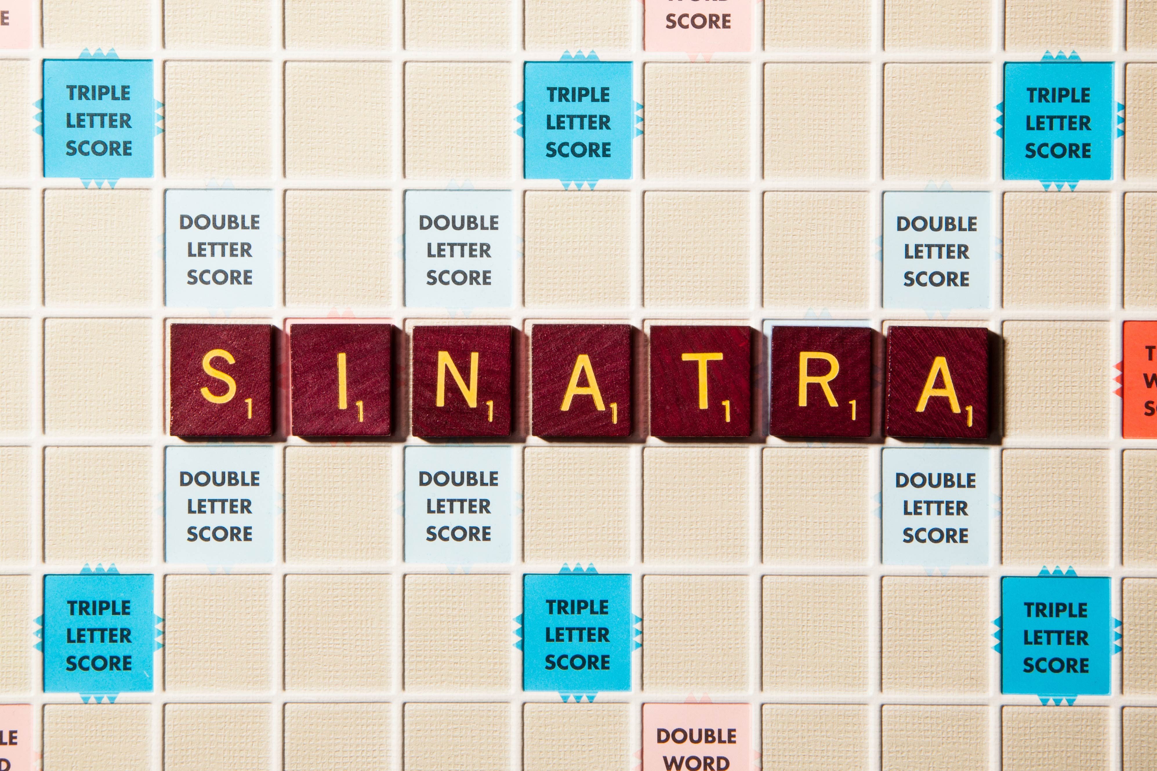 Brown Scrabble tiles spell "SINATRA" horizontally on a game board, surrounded by scoring squares like "DOUBLE LETTER SCORE" and "TRIPLE LETTER SCORE."