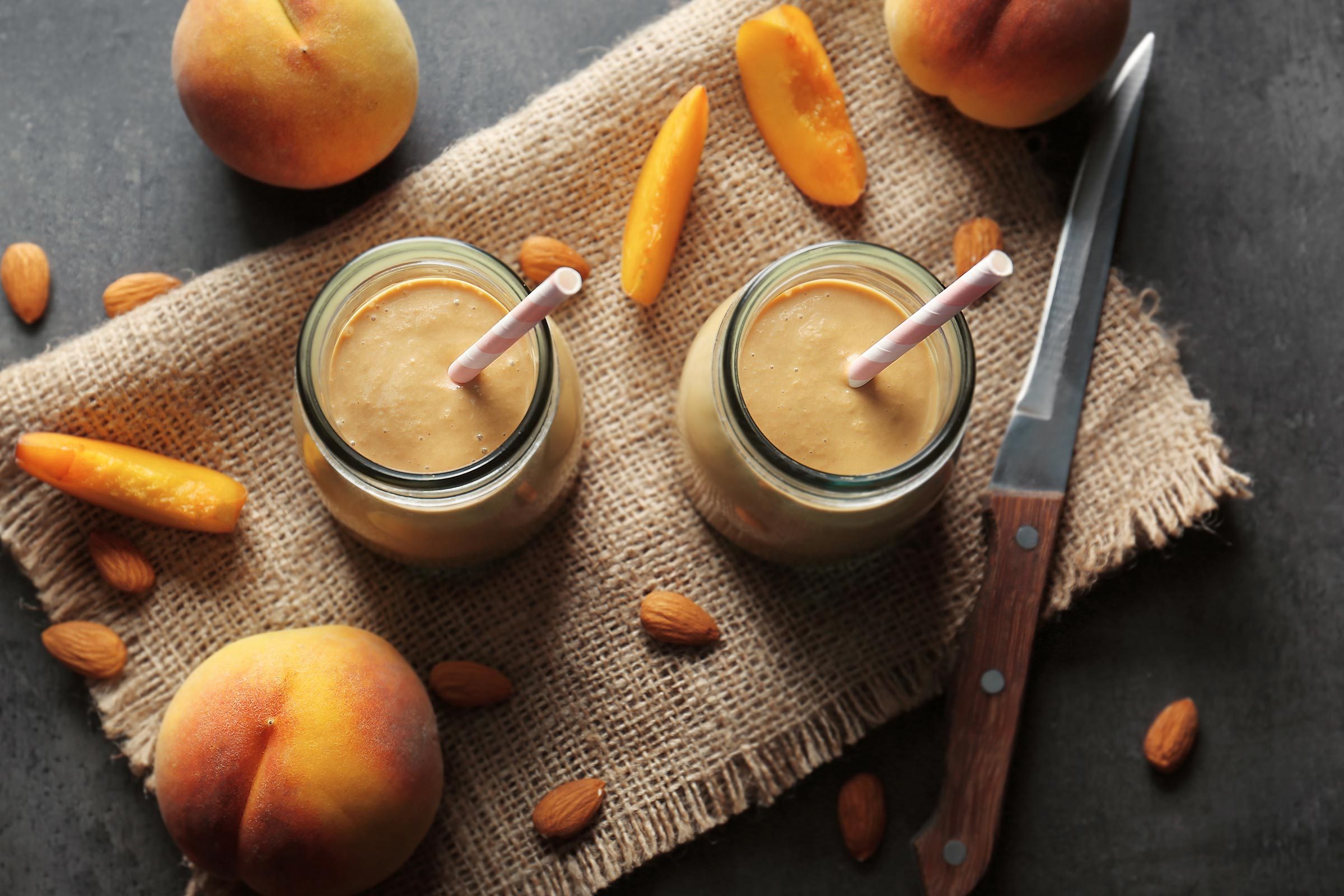 Two jars of peach smoothie with straws sit on burlap, surrounded by peaches, almonds, and a knife on a dark surface.