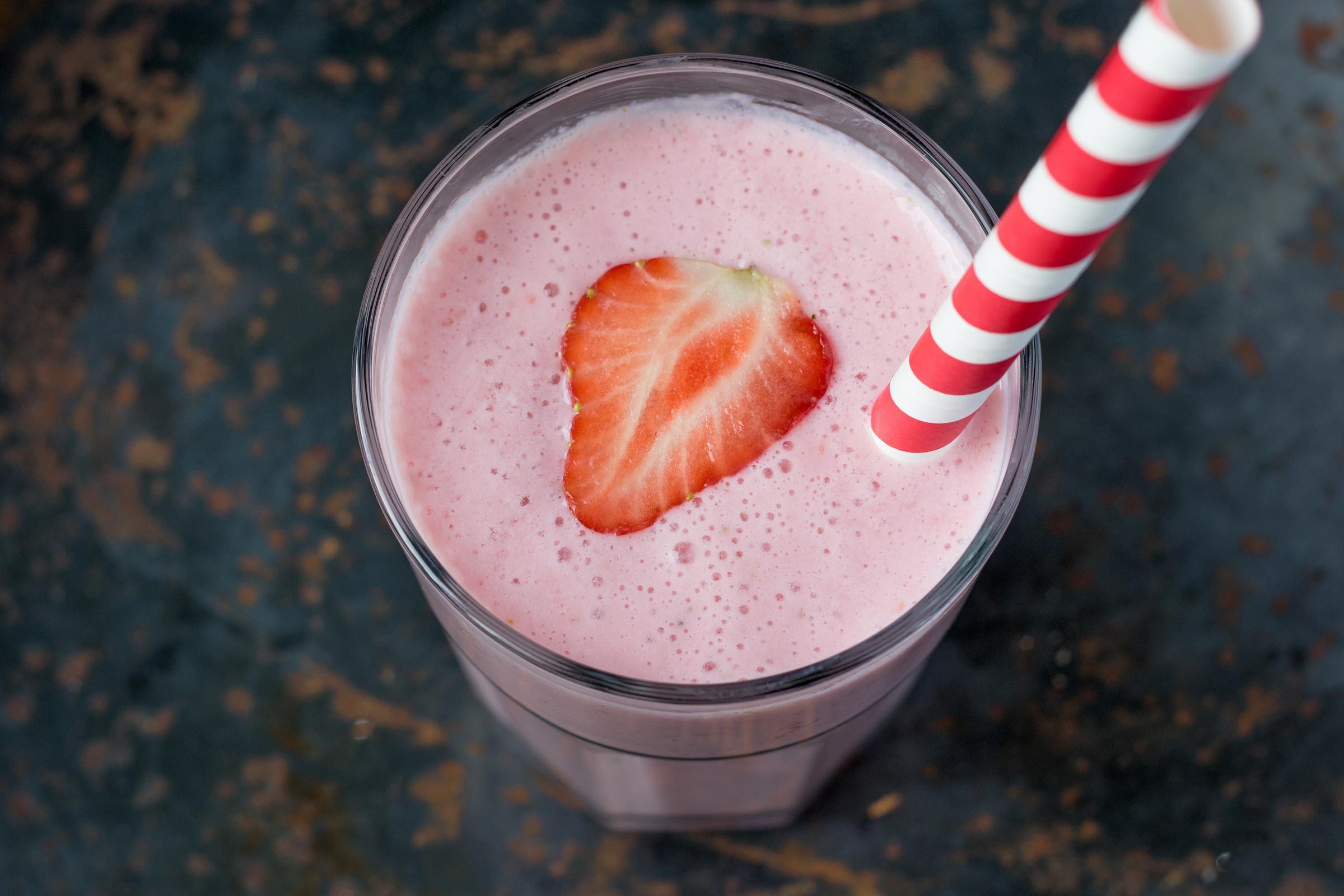 A pink smoothie with a strawberry slice and striped straw sits in a glass on a dark marbled surface.