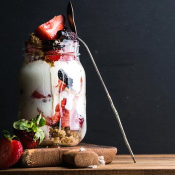 A jar filled with yogurt, strawberries, and granola stands on a wooden board, with a spoon inserted, against a dark background.