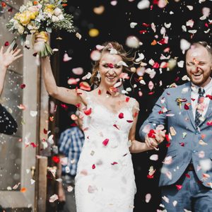 Newlyweds walk through a shower of colorful flower petals, the bride holding a bouquet, surrounded by joyous friends and photographers outside a building.