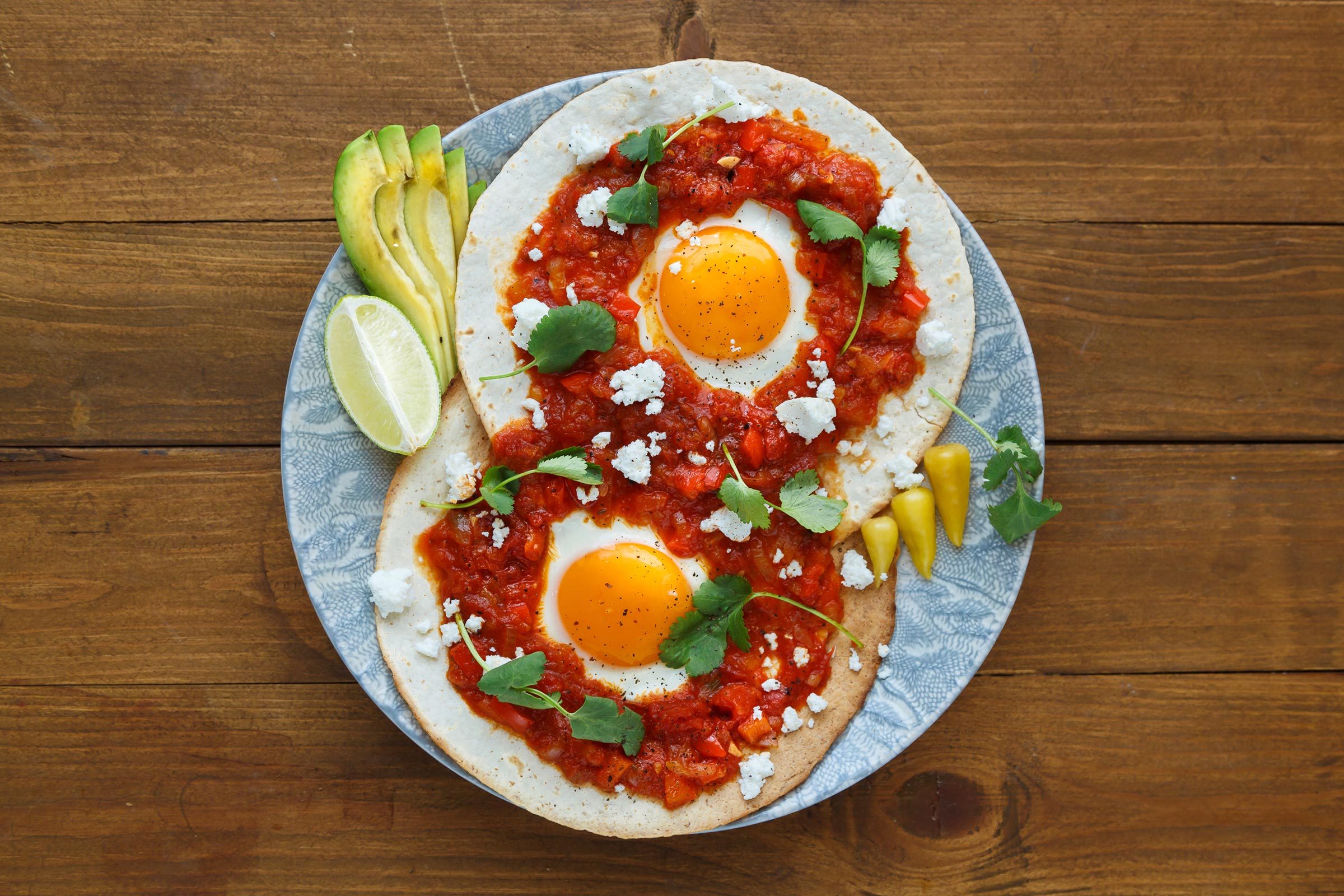 Eggs sit atop tortillas with salsa, garnished with cilantro and cheese; avocado, lime, and peppers on the side, on a wooden table.