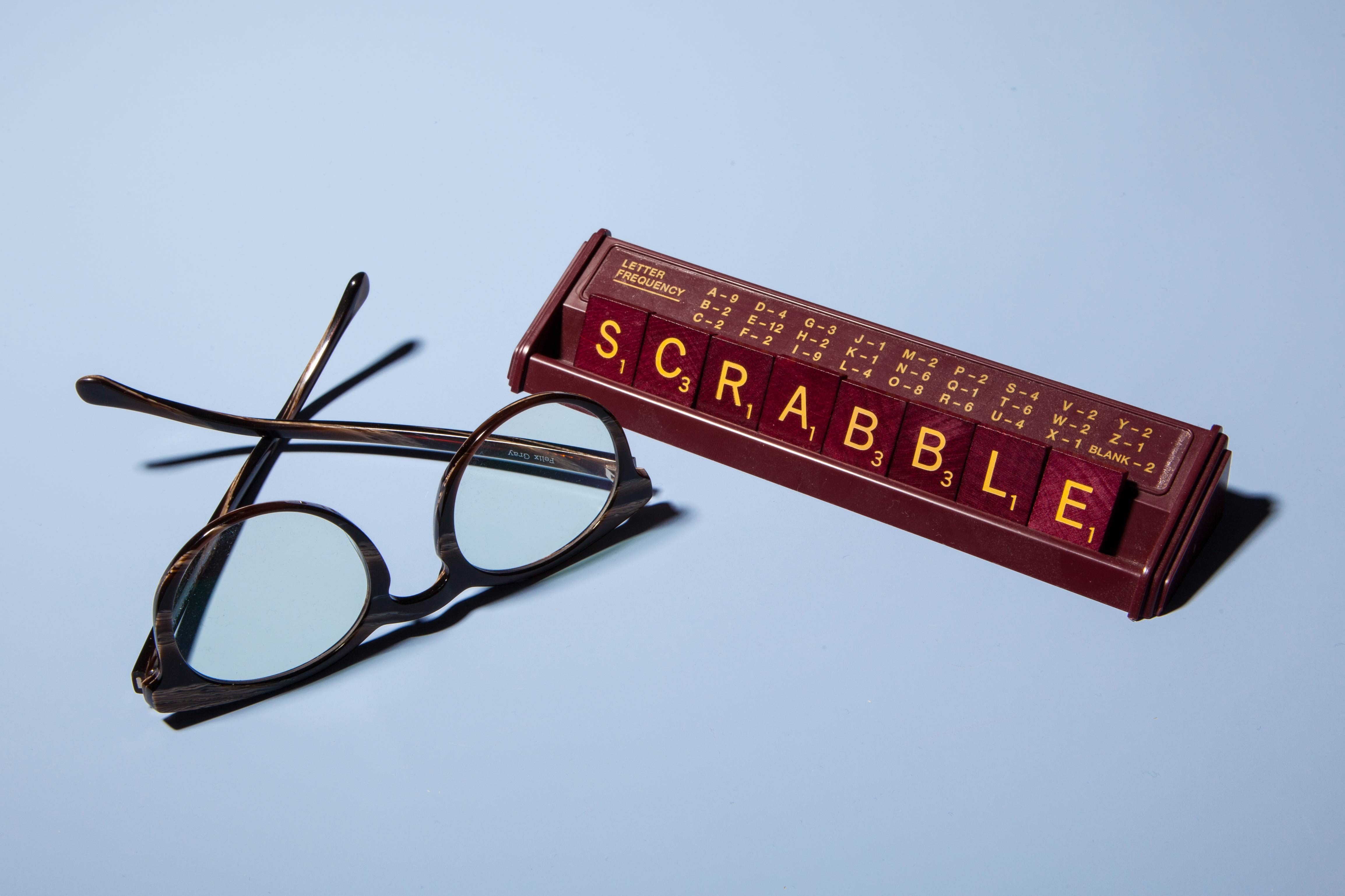 Glasses rest beside Scrabble tiles on a rack spelling "SCRABBLE," set against a light blue background.