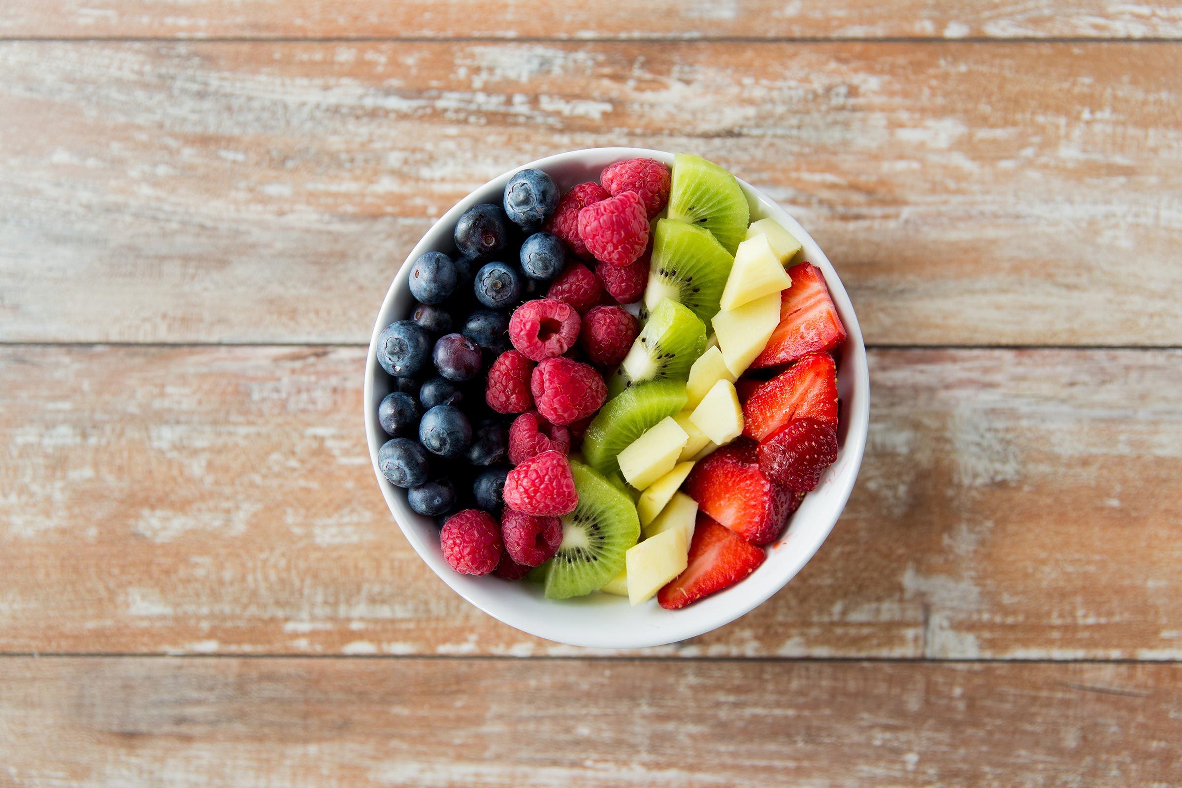 Bowl holds assorted berries, kiwi, and apple pieces, arranged neatly on a rustic wooden table surface.