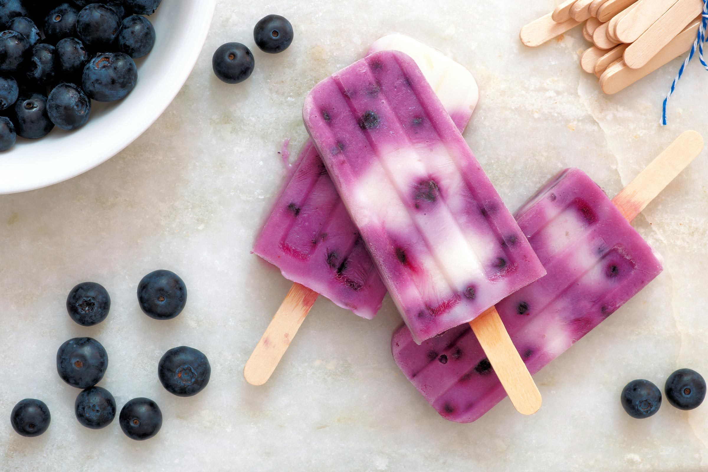 Purple and white popsicles rest on a marble surface, surrounded by fresh blueberries, near a bowl and stacked wooden sticks.